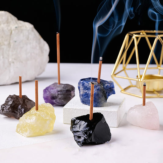 A collection of rough gemstone incense holders in various colors—black, yellow, blue, purple, and pink—each holding a burning incense stick with smoke gently rising. A geometric gold decor piece sits in the background, adding a touch of elegance.